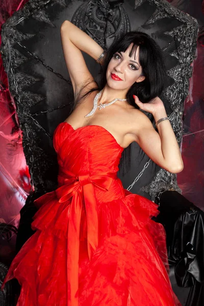 Beautiful vampire woman in red long dress near big black throne in the studio Royalty Free Stock Photos