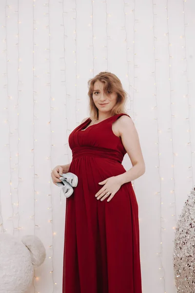 Jovem bela mulher grávida elegante em vestido de noite longo vermelho no fundo branco do quarto da criança — Fotografia de Stock