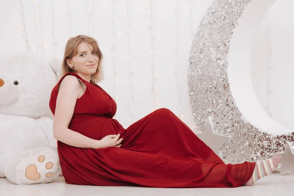 Νεαρή όμορφη κομψή έγκυος γυναίκα σε κόκκινο μακρύ βραδινό φόρεμα σε λευκό φόντο του παιδικού δωματίου — Φωτογραφία Αρχείου