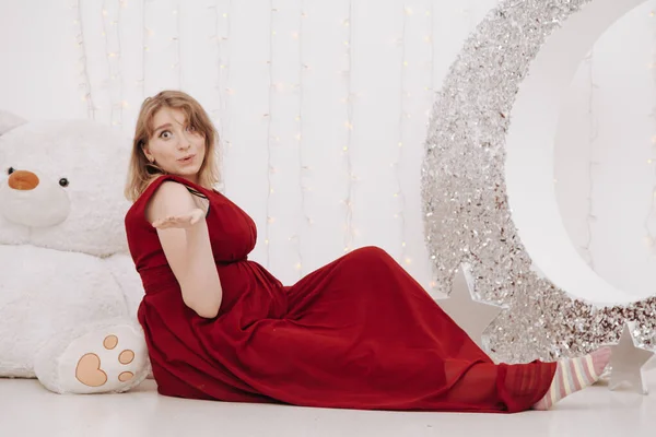 Jovem bela mulher grávida elegante em vestido de noite longo vermelho no fundo branco do quarto da criança — Fotografia de Stock