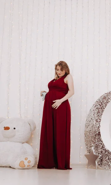 Young beautiful stylish pregnant woman in red long evening dress on white background of child room — Stock Photo, Image