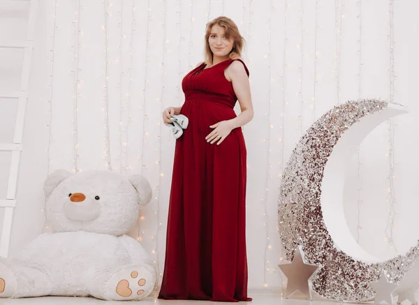 Ung vacker snygg gravid kvinna i röd lång kväll klänning på vit bakgrund av barnrum — Stockfoto