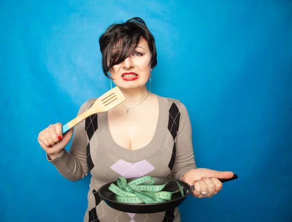 Una chica emocional de talla grande con un corte de pelo corto se para en un suéter y sostiene una sartén vacía y una cinta de centímetro, muestra que está a dieta y tratando de perder peso — Foto de Stock