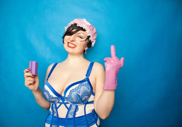 Adorable happy plus size short hair woman in blue white lingerie with a rubber glove and a sponge for washing dishes on a solid background in Studio — ストック写真