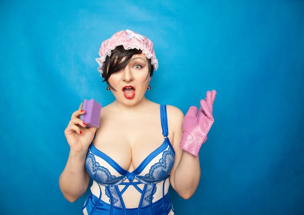 Adorable happy plus size short hair woman in blue white lingerie with a rubber glove and a sponge for washing dishes on a solid background in Studio — ストック写真