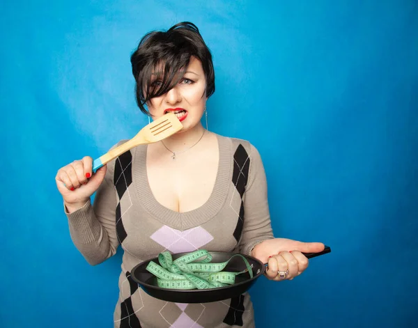 Una chica emocional de talla grande con un corte de pelo corto se para en un suéter y sostiene una sartén vacía y una cinta de centímetro, muestra que está a dieta y tratando de perder peso — Foto de Stock