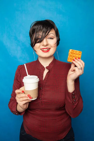 Alegre mulher plus size com cabelo curto em roupas de negócios descansando e bebendo café com uma palha com waffle vienense como lanche em um fundo de estúdio azul . — Fotografia de Stock