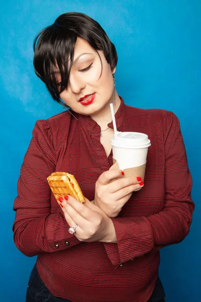 Alegre mujer de tamaño grande con pelo corto en ropa de negocios descansando y bebiendo café con una paja con gofre vienés como merienda en un fondo de estudio azul . — Foto de Stock