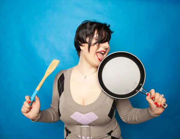 Front view portrait of a young happy caucasian female dressed in sweater, holding a frying pan in front of her, on blue studio background — Stock Photo, Image