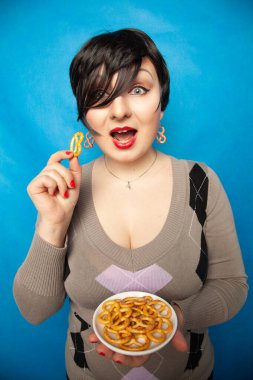 Süveter giyen çekici ve cüsseli bir kız bir tabak tuzlu krakerle stüdyoda mavi bir arka planda atıştırmaktan hoşlanıyor.