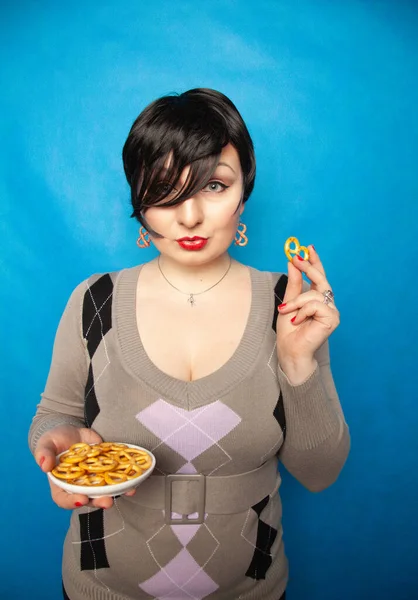 Encantadora chica de tamaño grande en un suéter se encuentra con un plato de pretzels salados y disfruta de un aperitivo sobre un fondo azul en el estudio — Foto de Stock