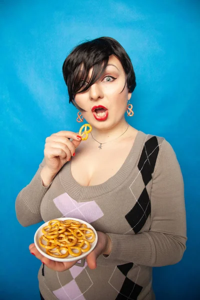 Encantadora chica de tamaño grande en un suéter se encuentra con un plato de pretzels salados y disfruta de un aperitivo sobre un fondo azul en el estudio — Foto de Stock