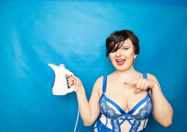 Cute adult girl with plus size figure stands in beautiful lingerie with a small travel steamer in her hand on a blue background in the Studio — Stock Photo, Image