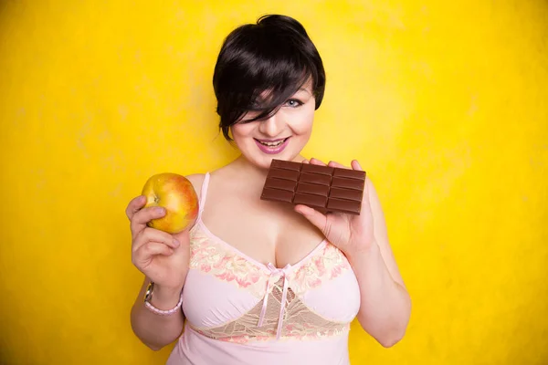 Uma menina atraente plus size pesa suas opções e decidir comer chocolate ou uma maçã no fundo do estúdio amarelo brilhante — Fotografia de Stock