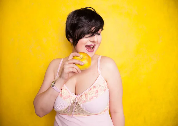 Pretty woman with overweight bite the Apple and lose weight on yellow studio background — Stock Photo, Image