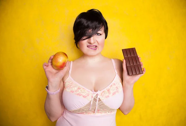 Atraktivní plus velikost dívka váží své možnosti a rozhodnout se jíst čokoládu nebo jablko na jasně žlutém pozadí studia — Stock fotografie