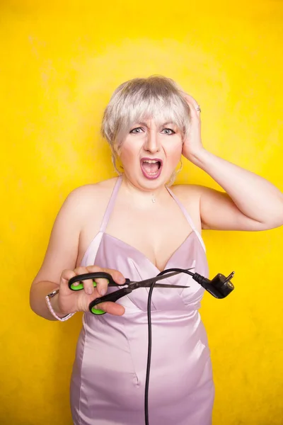 Stomme persoon snijdt elektrische draad door met een schaar. dom blond in roze jurk speelt slecht met elektriciteit op gele studio achtergrond — Stockfoto