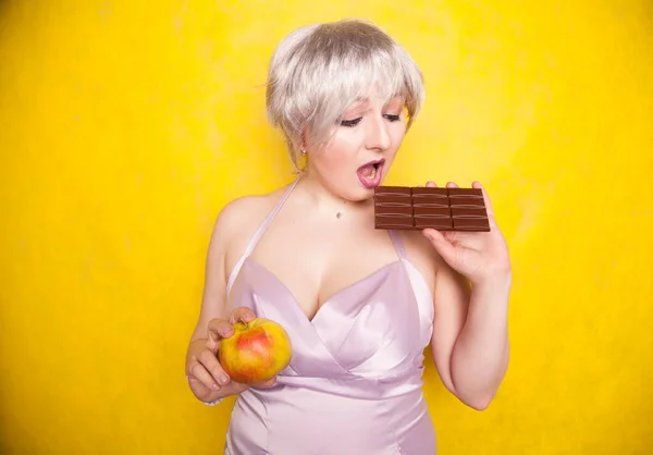 Doce alegre menina curvácea com um corte de cabelo curto fica em um vestido de seda noite delicada e compara má e boa comida, decidindo que saúde ela quer para si mesma — Fotografia de Stock