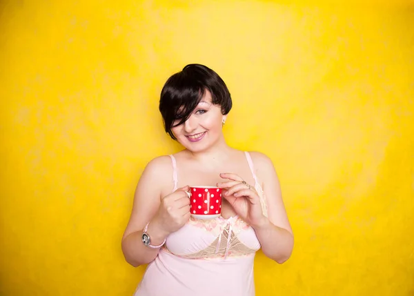 Mujer de tamaño grande con vestido rosa sosteniendo un café de cerámica de lunares rojos o una taza de té sobre un fondo amarillo brillante — Foto de Stock