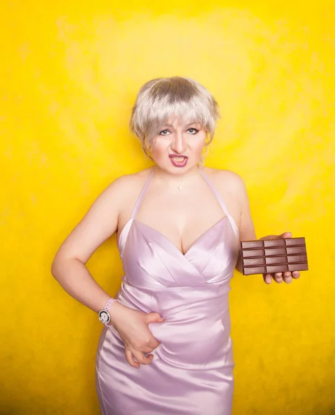 Kobiece palce mierzą jej tłuszcz brzucha, a ona trzyma czekoladę na żółtym stałym tle studio. — Zdjęcie stockowe