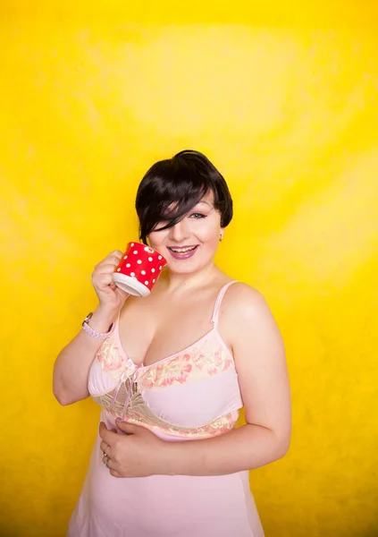 Além de mulher de tamanho usando vestido rosa segurando um café vermelho bolinha cerâmica ou xícara de chá em um fundo amarelo brilhante — Fotografia de Stock