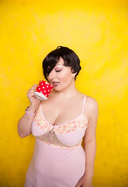 Plus ukuran wanita mengenakan gaun merah muda memegang merah polka dot kopi keramik atau cangkir teh pada latar belakang kuning cerah — Stok Foto