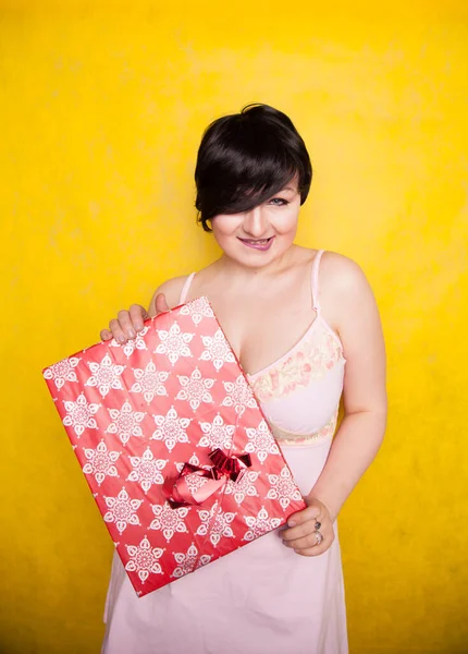 Happy Female holding in hands big gift with ribbon. Adult girl with red present on yellow studio background. — ストック写真