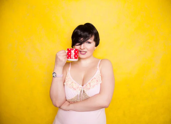 Plus sized woman wearing pink dress holding a red polka dot ceramic coffee or tea cup on a bright yellow background — Stock Photo, Image