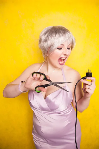 Stupid person cuts electric wire with scissors. silly blonde in pink dress plays bad with electricity on yellow studio background — ストック写真