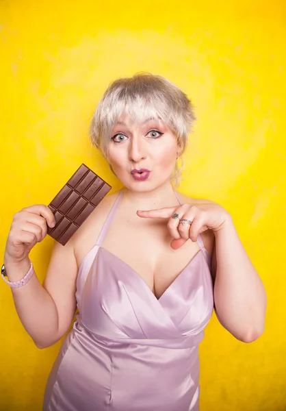 Hezká blondýnka krátké vlasy žena s nadváhou držení v ruce velký čokoládový prut na žluté světlé pozadí studia — Stock fotografie
