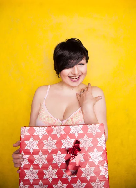Happy Female holding in hands big gift with ribbon. Adult girl with red present on yellow studio background. — Stock Photo, Image