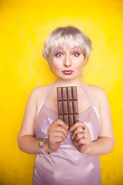 Hezká blondýnka krátké vlasy žena s nadváhou držení v ruce velký čokoládový prut na žluté světlé pozadí studia — Stock fotografie