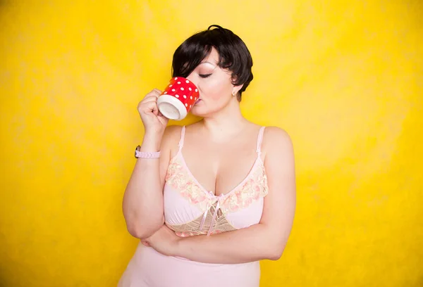 Plus sized woman wearing pink dress holding a red polka dot ceramic coffee or tea cup on a bright yellow background — ストック写真