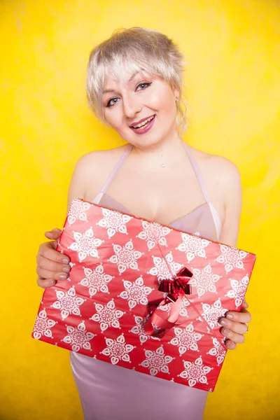 Sweet joyous curvaceous girl with a short haircut and plus size body stands in a delicate evening lilac silk dress and holds a large bright red box with a gift for the holiday — Stock Photo, Image