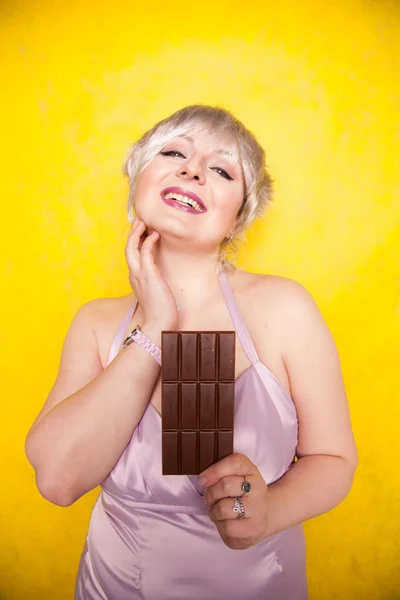 Bonita loira mulher cabelo curto com o excesso de peso segurando em suas mãos uma grande barra de chocolate no amarelo brilhante estúdio fundo — Fotografia de Stock