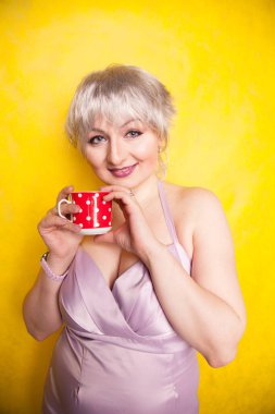 Kısa saçlı güzel, kıvrımlı sarışın kadın sarı stüdyo arka planında kırmızı benekli seramik bardaktan çay içmeyi seviyor.