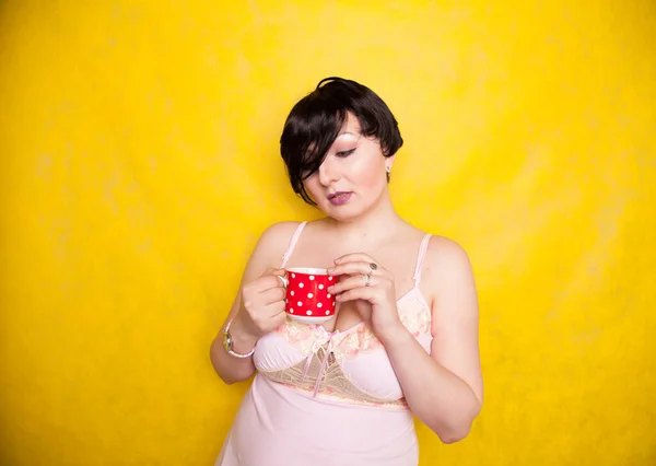 Plus sized woman wearing pink dress holding a red polka dot ceramic coffee or tea cup on a bright yellow background — Stock Photo, Image