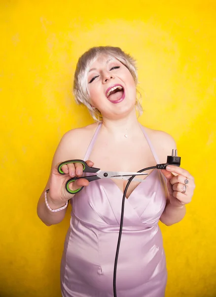 Stupid person cuts electric wire with scissors. silly blonde in pink dress plays bad with electricity on yellow studio background — Stock Photo, Image