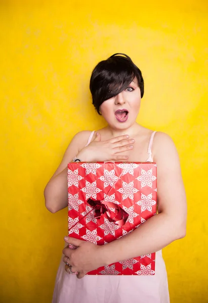 Happy Female holding in hands big gift with ribbon. Adult girl with red present on yellow studio background. — ストック写真