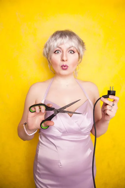 Stomme persoon snijdt elektrische draad door met een schaar. dom blond in roze jurk speelt slecht met elektriciteit op gele studio achtergrond — Stockfoto