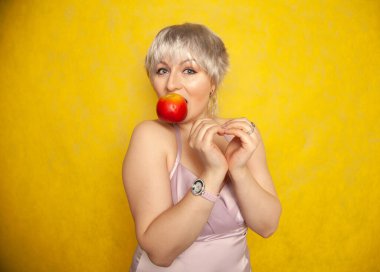 Çekici, komik ve kısa saçlı bir kadın elleriyle kalp sembolünü gösteriyor ve ağzında kırmızı bir elma tutuyor..