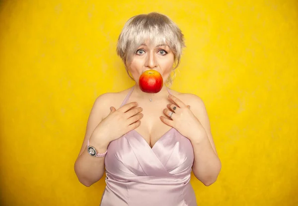 Okouzlující legrační plus velikost žena s krátkými vlasy ukazuje symbol srdce s rukama a drží červené jablko v ústech. — Stock fotografie
