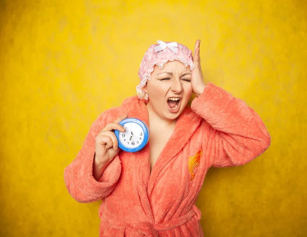 Girl in a Terry robe and with a shower cap on her head stands with an alarm clock in her hand and shows negative emotions on a yellow background in the Studio — Stockfoto