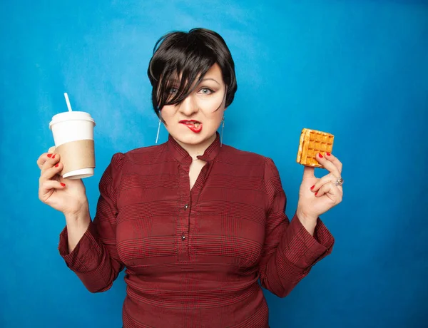 Joyful plus size woman with short hair in business clothes resting and drinking coffee with a straw with Viennese waffle as snack on a blue studio background. — ストック写真