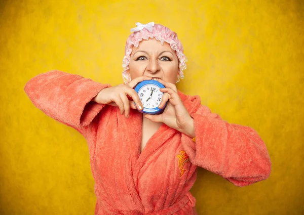 Girl in a Terry robe and with a shower cap on her head stands with an alarm clock in her hand and shows negative emotions on a yellow background in the Studio — Stockfoto