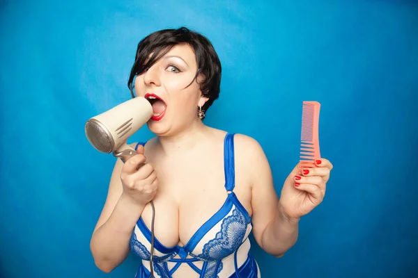 Cute plus size girl with short black hair stands in lingerie and sing with a travel small hair dryer in hand on blue studio background alone — Stock Photo, Image
