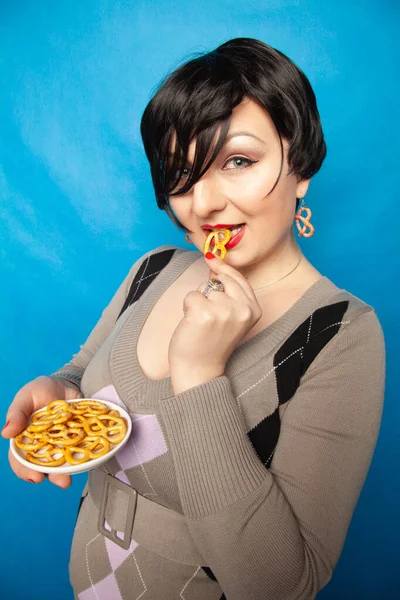 Encantadora chica de tamaño grande en un suéter se encuentra con un plato de pretzels salados y disfruta de un aperitivo sobre un fondo azul en el estudio — Foto de Stock