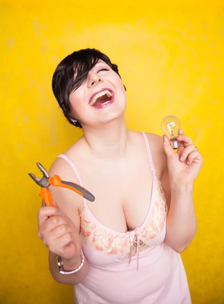 Cute funny girl in lingerie stands with pliers and a light bulb and does not know what to do with them, how to make repairs — Stock Photo, Image