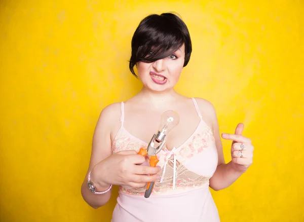 Cute funny girl in lingerie stands with pliers and a light bulb and does not know what to do with them, how to make repairs — Stock Photo, Image