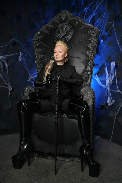 Foto de uma fêmea bruxa rainha segurando pássaro e sentado em um trono negro assustador gótico — Fotografia de Stock